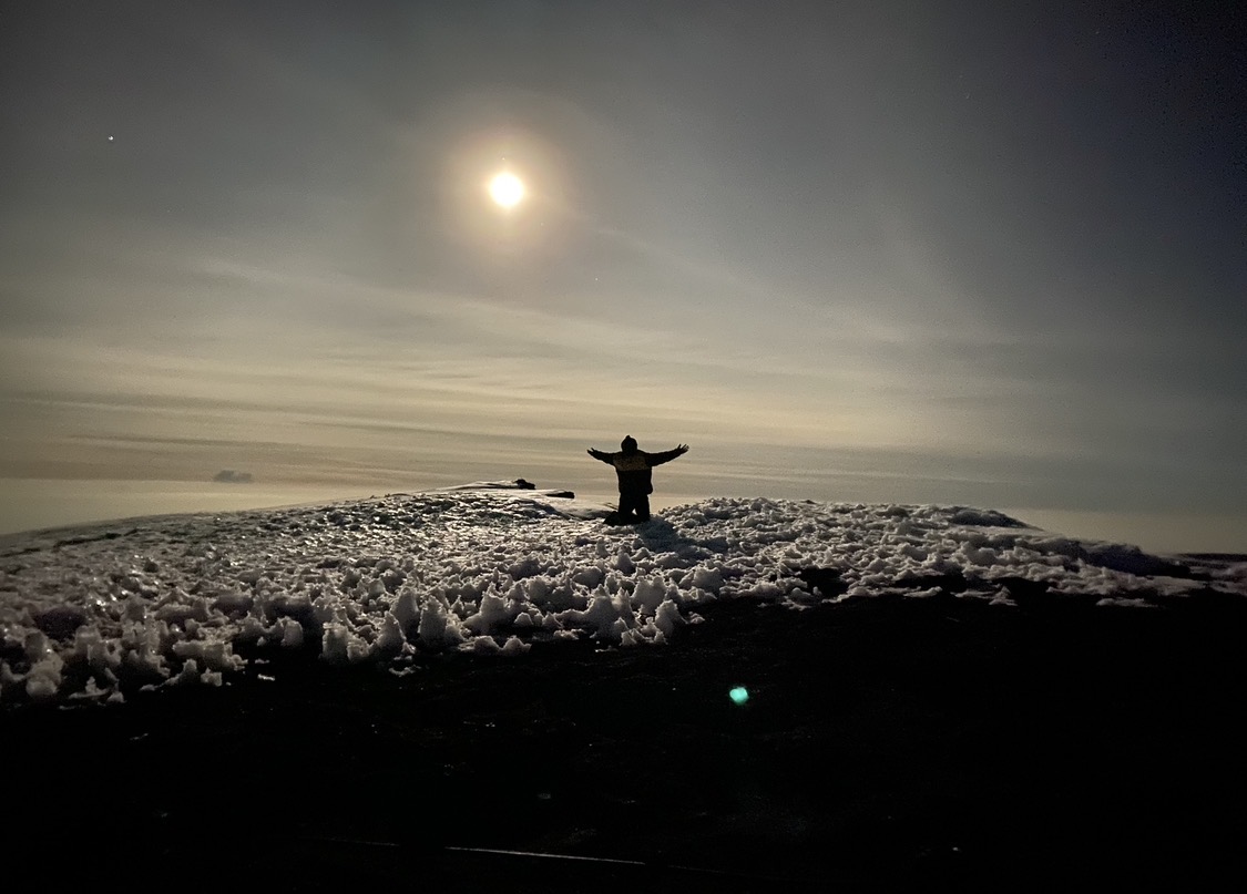 A lone climber stands on a snowy summit with arms raised beneath a moonlit sky.
